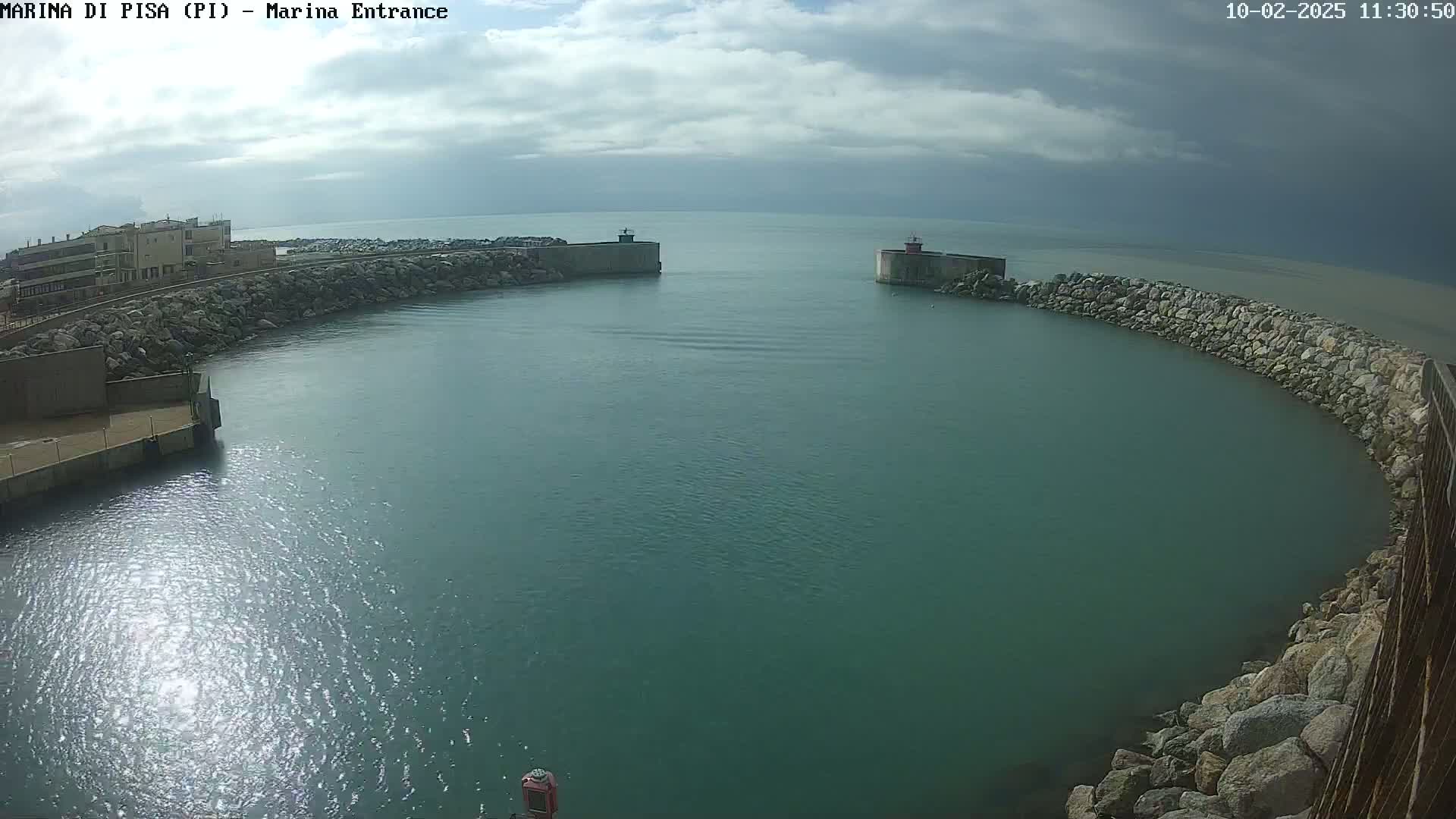 A partly cloudy day shows a calm harbor entrance with breakwaters and buildings visible in the distance.