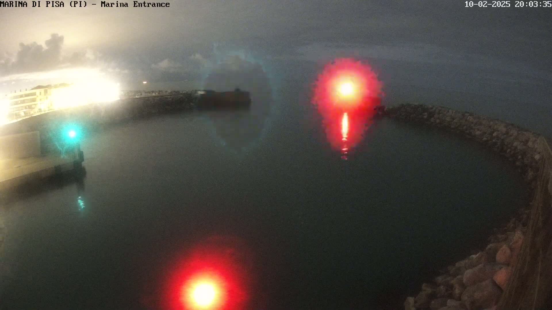A nighttime aerial view of a harbor entrance with red and green navigation lights and a bright red light seemingly emanating from a buoy or flare.