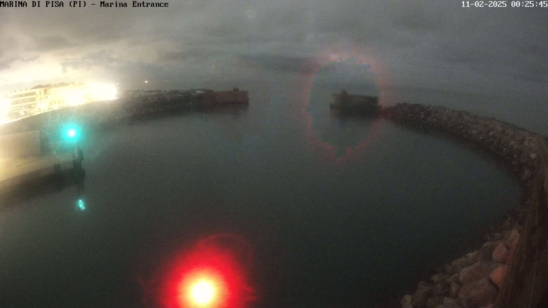 A nighttime, hazy view of a harbor entrance shows dark water enclosed by a rocky wall, with buildings and lights visible in the background.