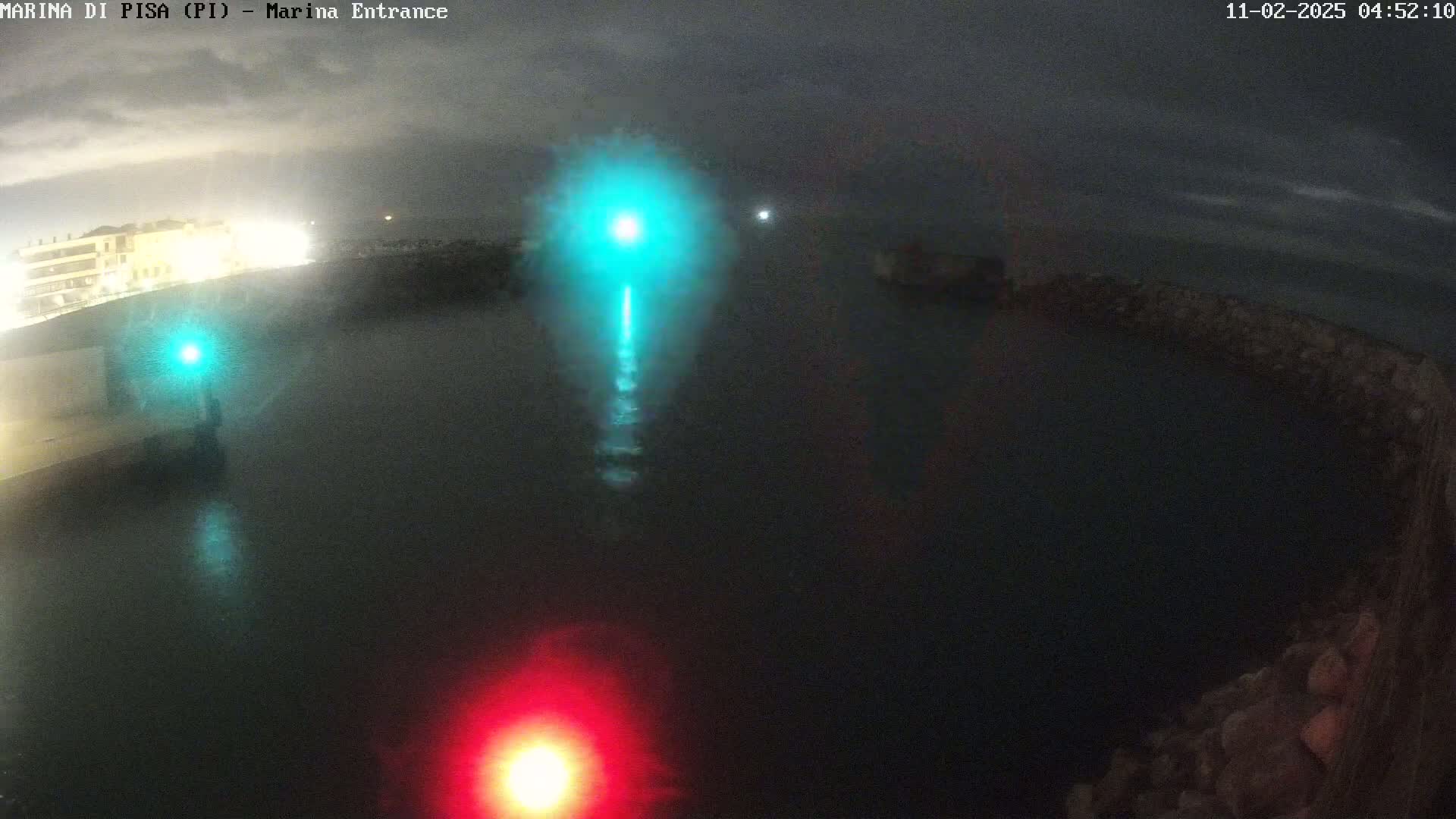 A nighttime view of a dark harbor with a bright teal light and a red light reflecting on the water under an overcast sky.