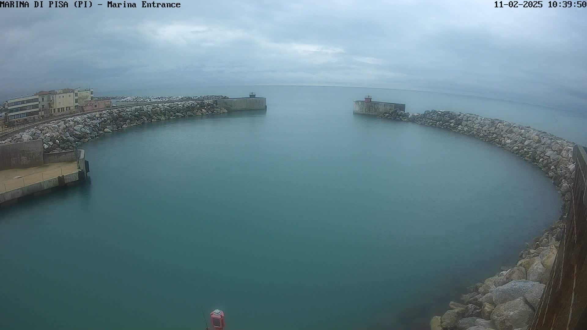 A harbor entrance with calm, teal water under an overcast sky is bordered by rock walls and a building.