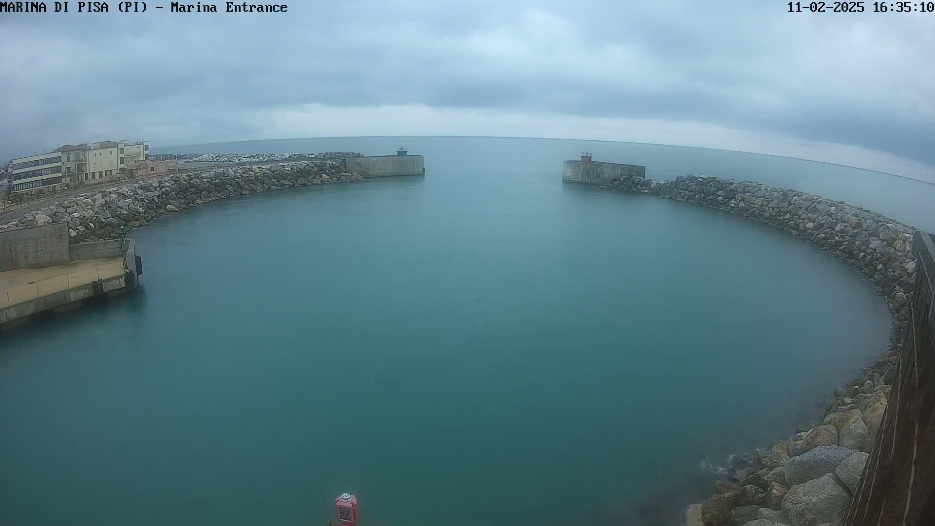 A harbor entrance with teal water is seen under a cloudy sky, bordered by rock walls and a building in the distance.