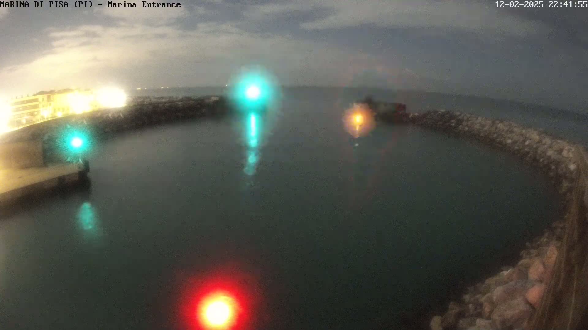 A nighttime view of a harbor entrance with calm, dark water, illuminated by several bright lights, including a green and a red light.