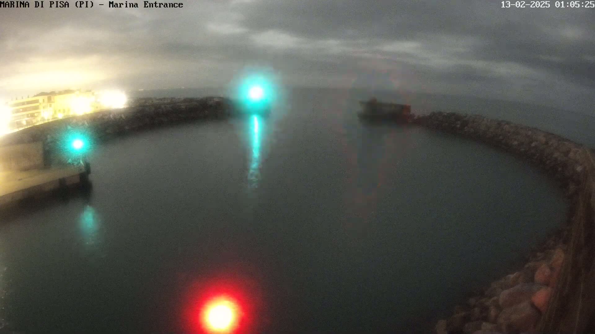 A nighttime, overcast view of a harbor entrance with dimly lit buildings and harbor walls, illuminated by a few lights reflecting in dark water.