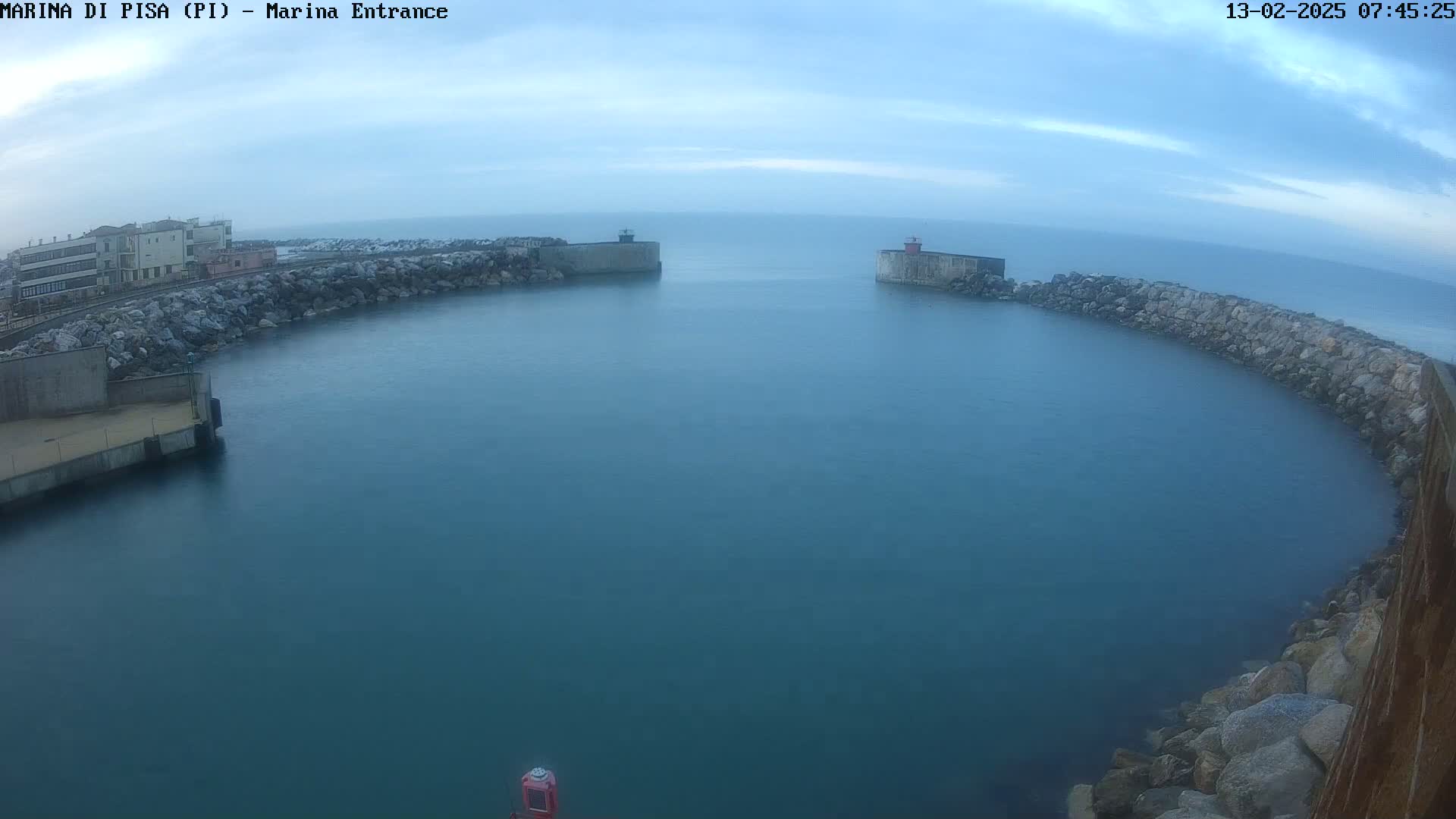 A calm, blue harbor is partially enclosed by stone walls and buildings under a mostly cloudy sky.