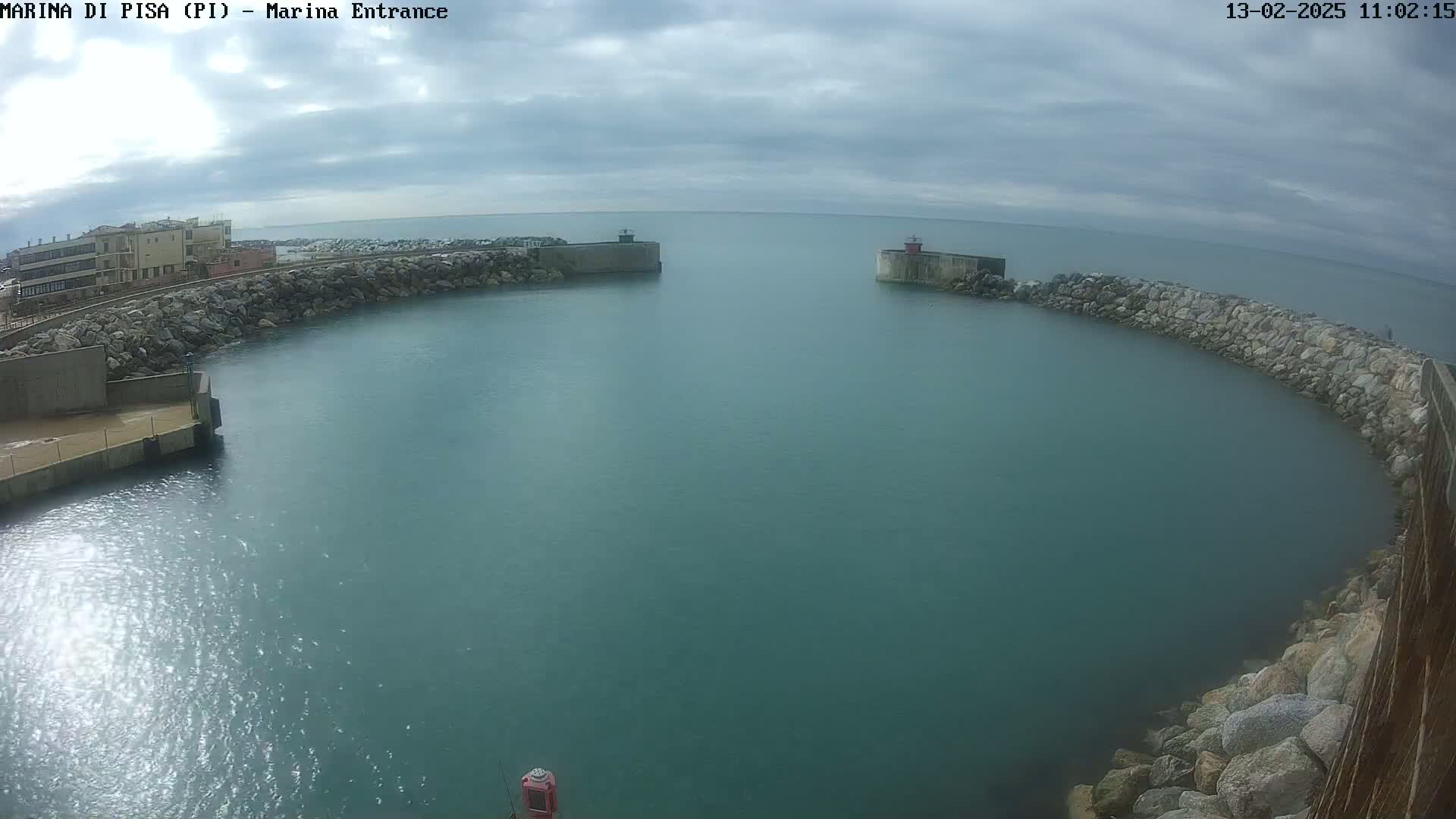 A harbor entrance with calm, teal water is framed by rock walls, under a cloudy sky.