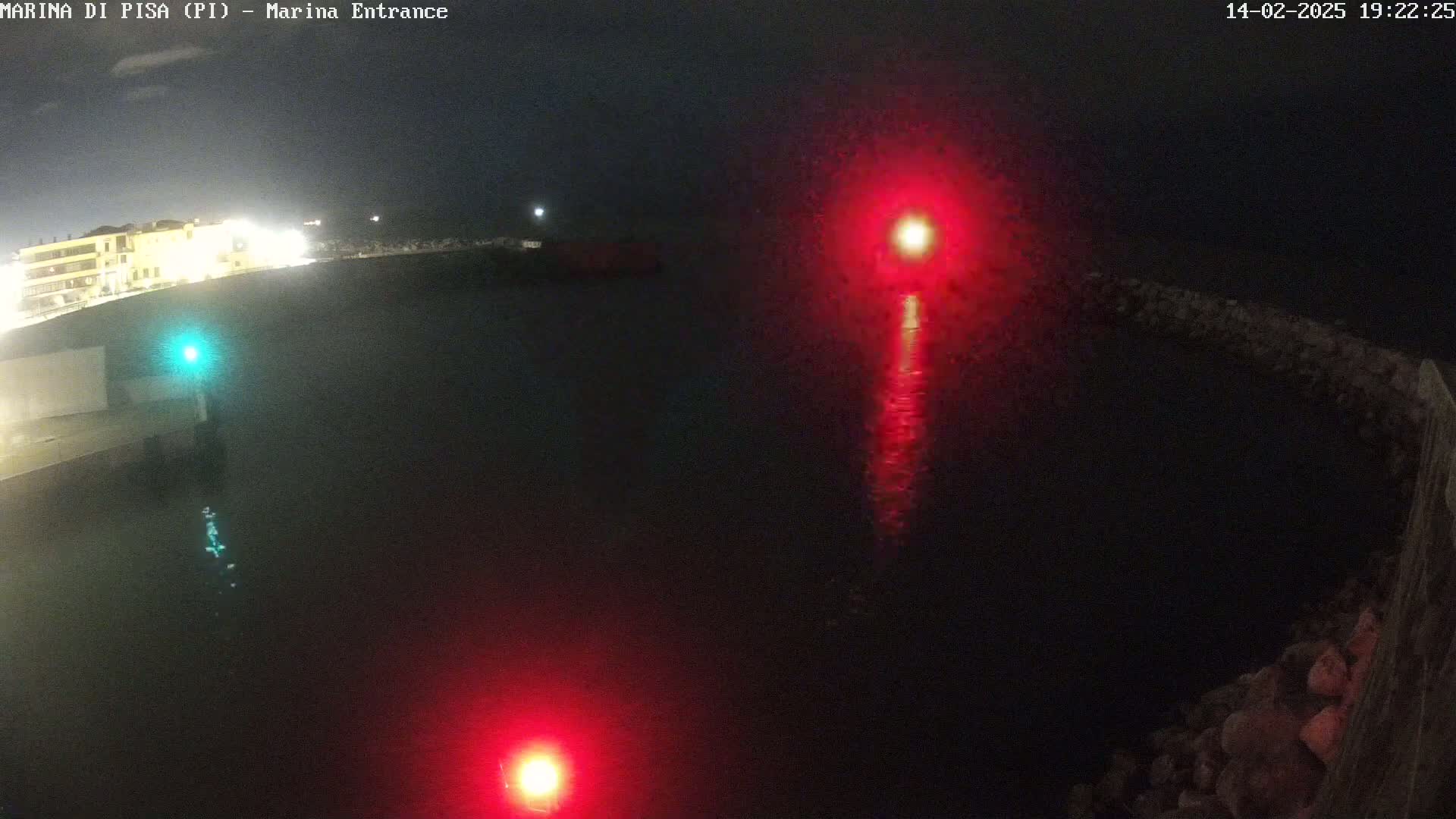 At night, a dark body of water reflects red and green lights from navigational markers near a shoreline with buildings.