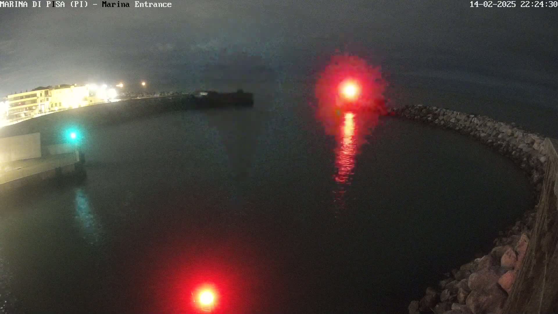 A nighttime aerial view shows a dark body of water enclosed by a curved rock wall, with red and green navigational lights illuminating the water.