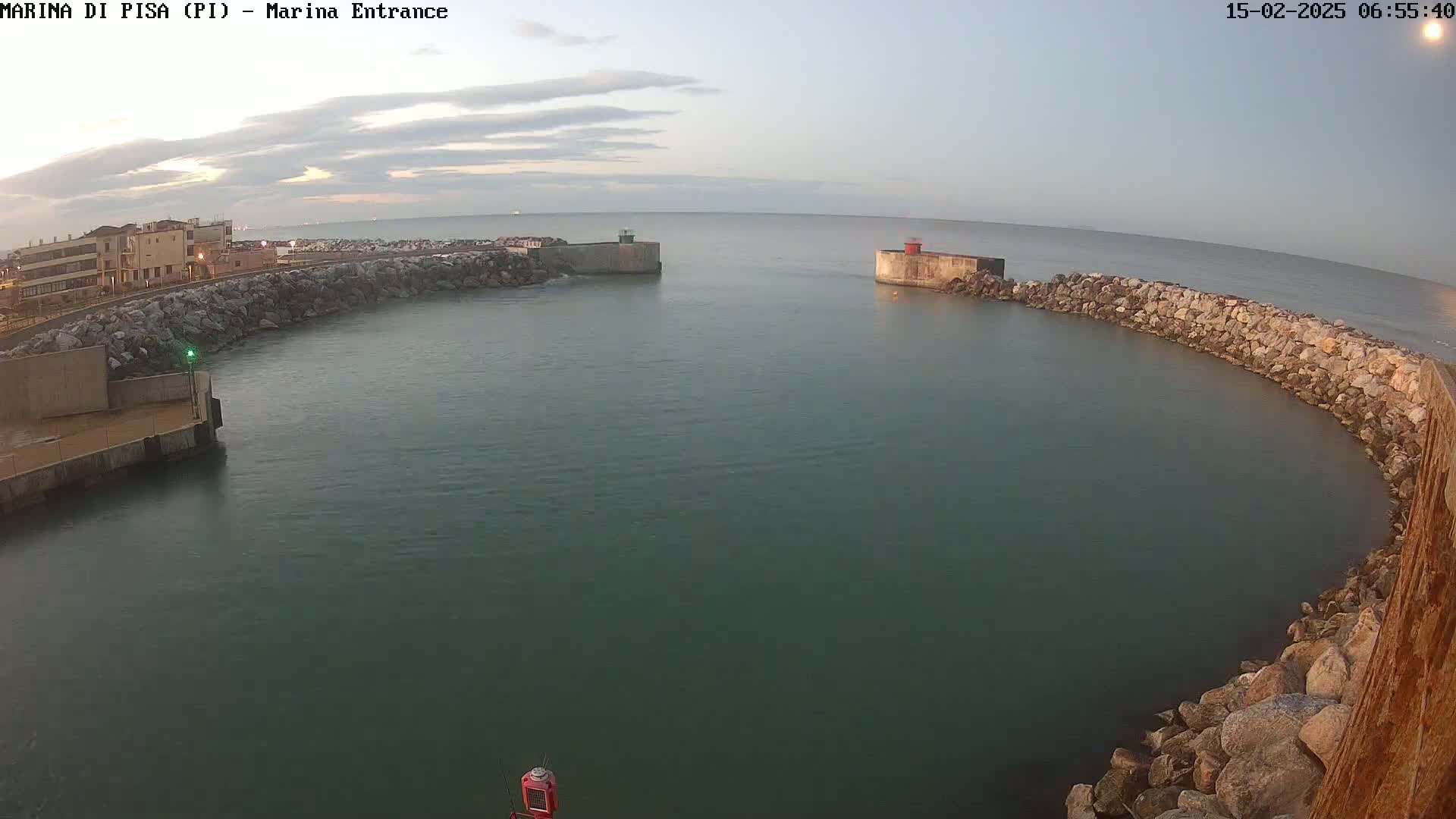 A calm, greenish-blue harbor is enclosed by rock walls, with a small red structure visible at the far end under a mostly clear sky.