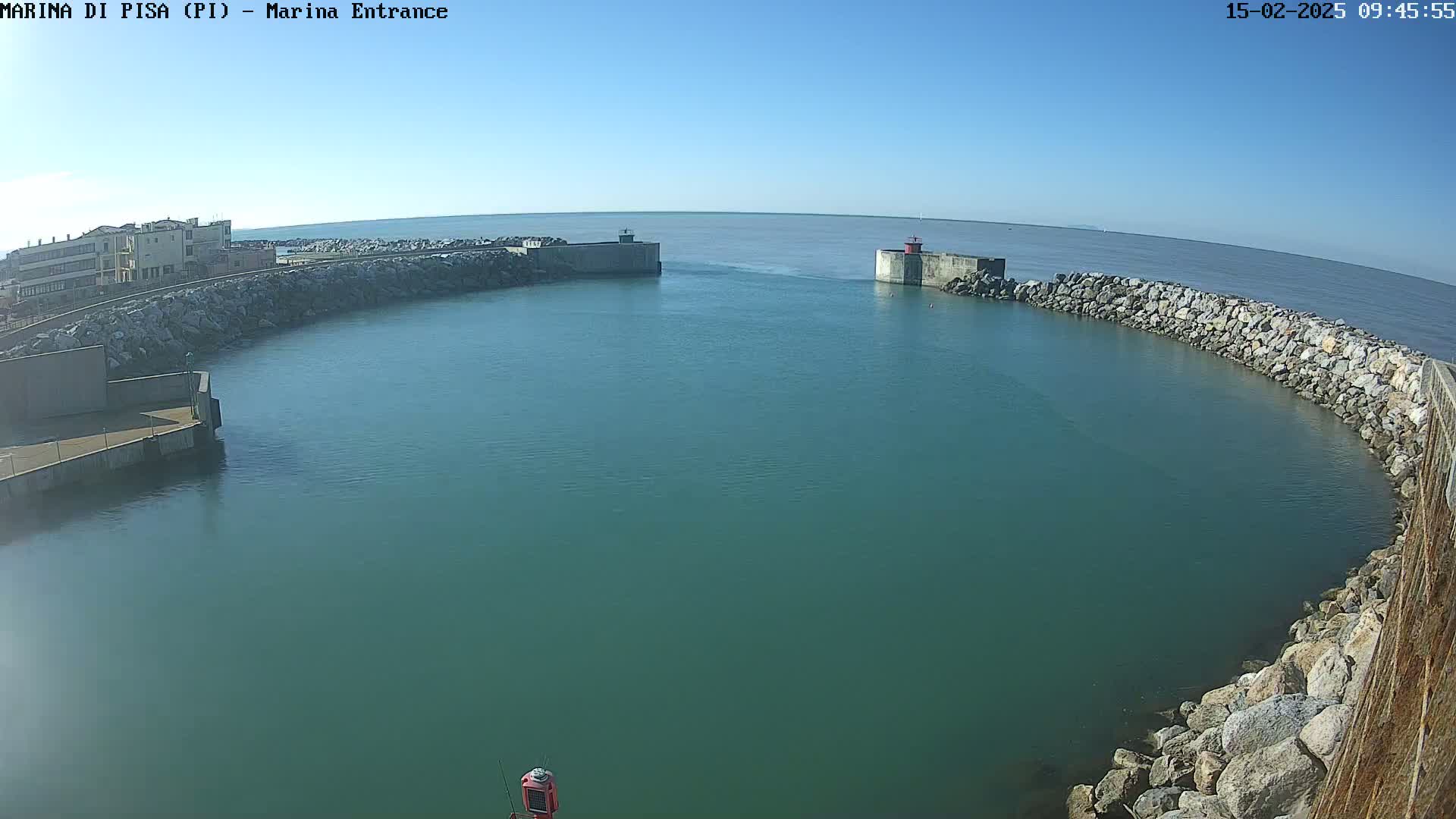 A calm, clear day shows a marina entrance with teal-colored water, rock walls, and a small building in the background.