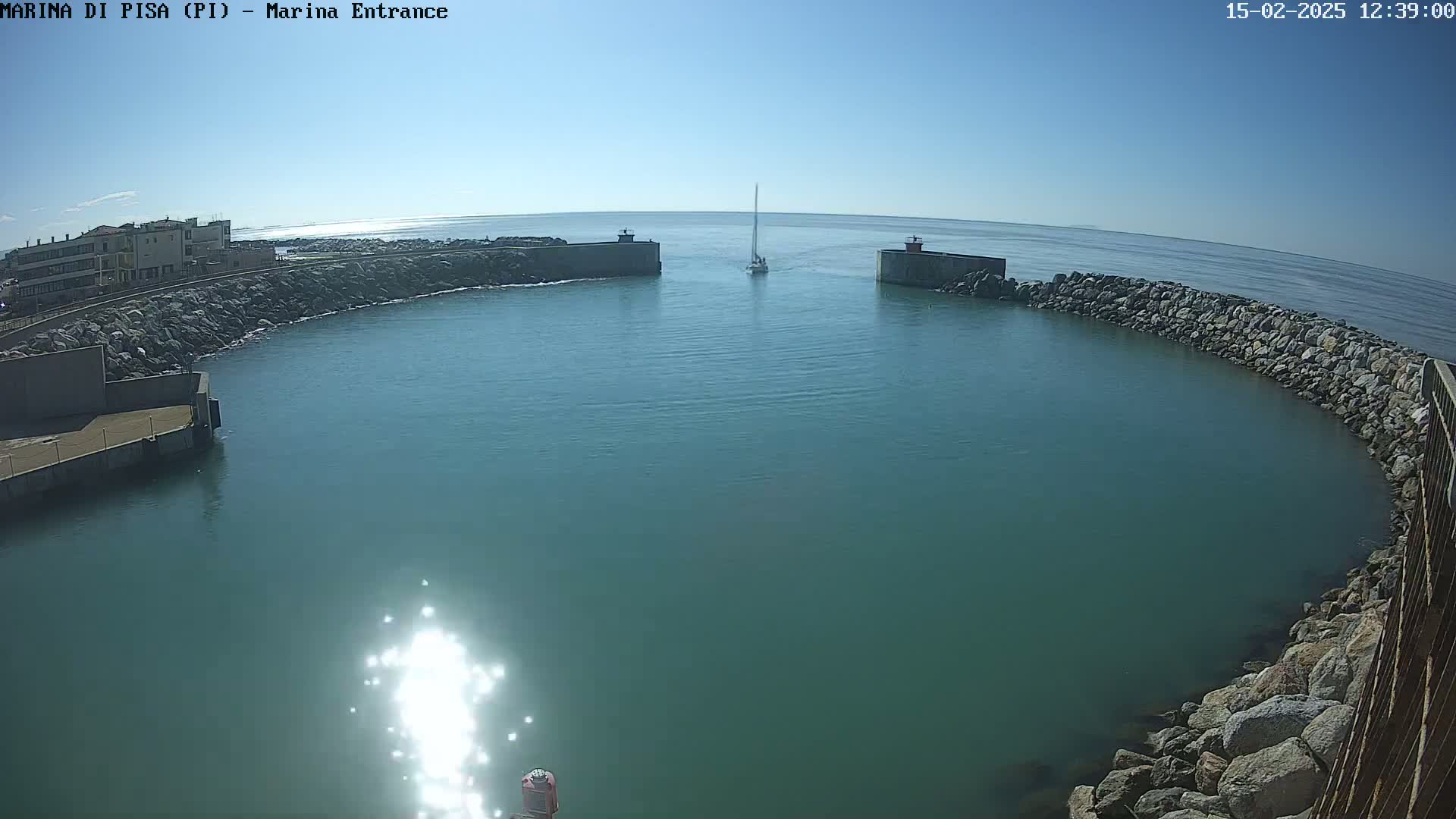A sailboat enters a harbor on a sunny day, passing between two rock jetties.