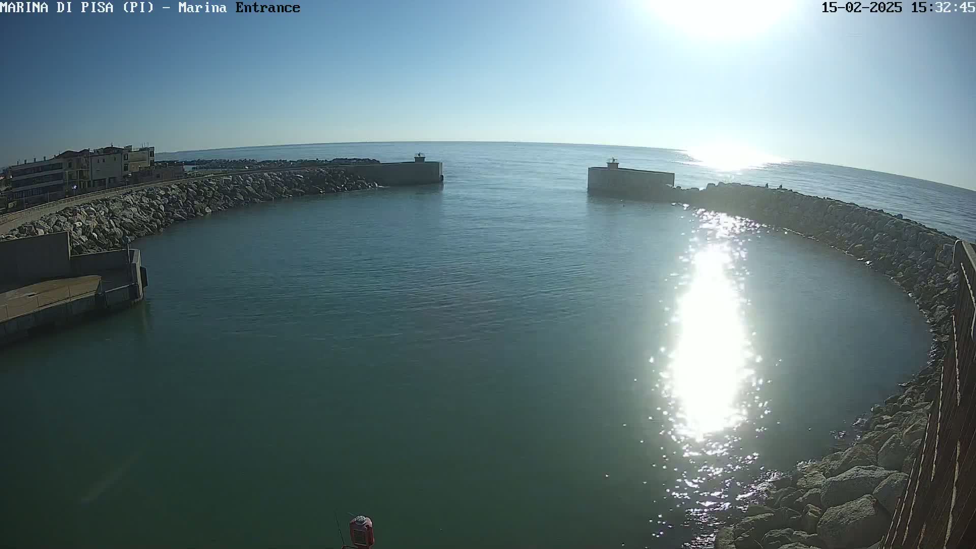 A calm, sunny day reveals a marina entrance with stone walls enclosing a tranquil body of water, reflecting sunlight.
