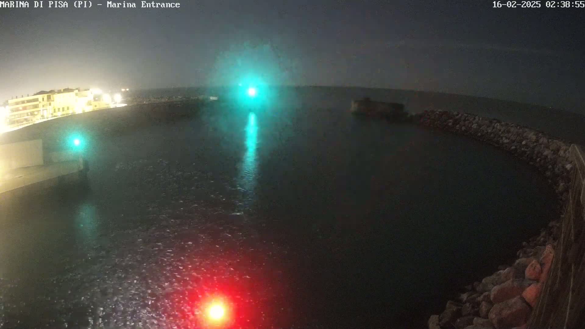 A nighttime view of a harbor entrance with a rocky breakwater, illuminated by lights, under rainy conditions.