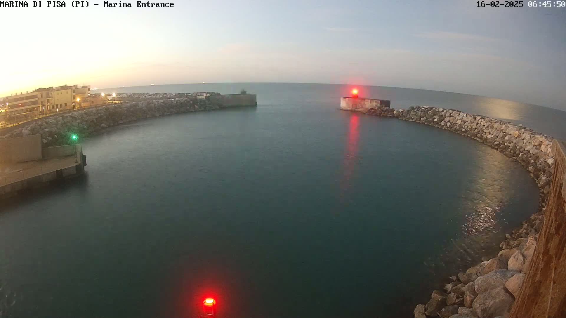 A calm, dark-blue body of water is enclosed by a curved stone wall at dawn, with buildings visible in the distance and a red light on a structure in the water.