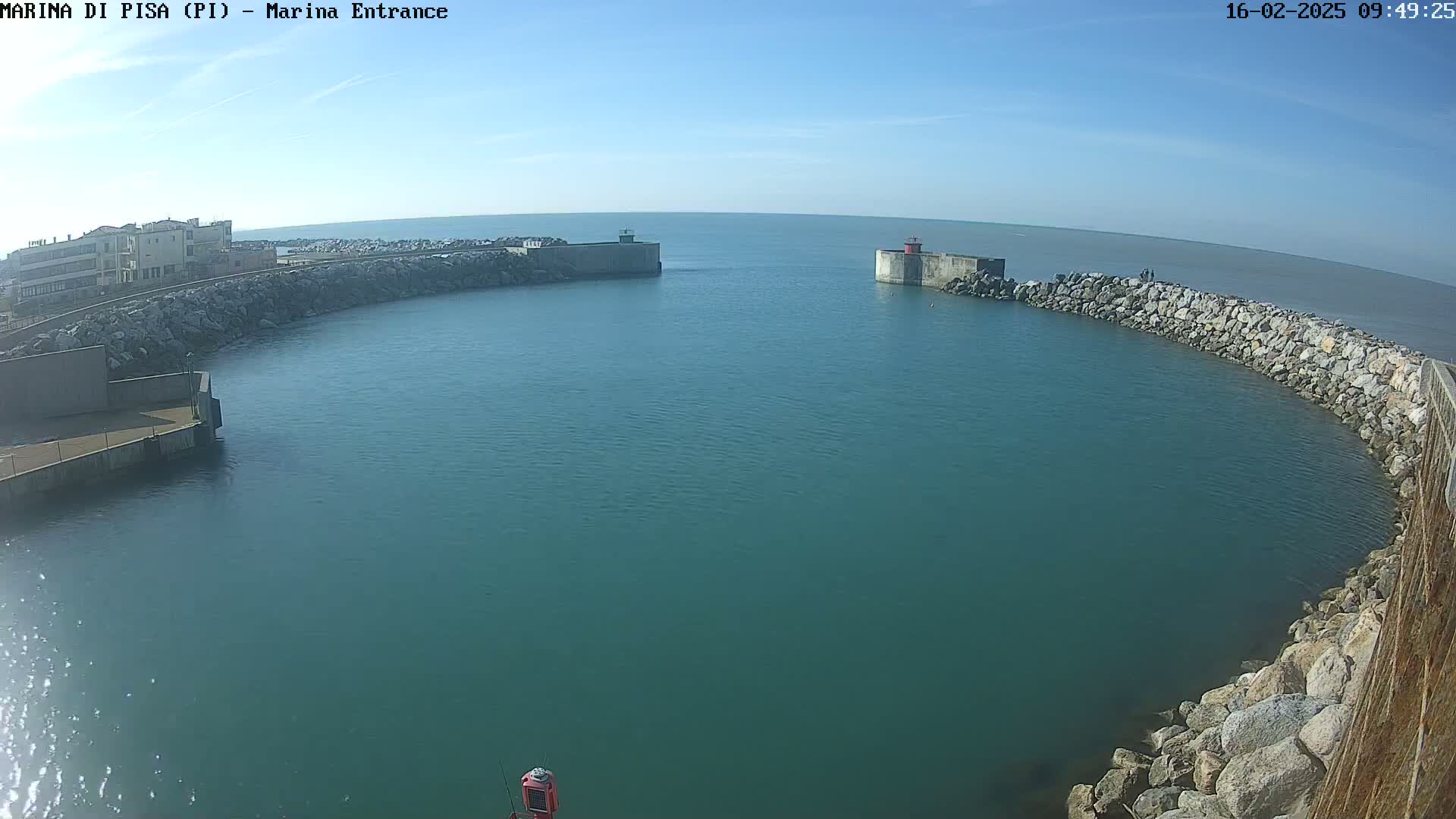 A calm, sunny day reveals a harbor entrance with a small building, rocky walls, and a mostly clear teal sea.