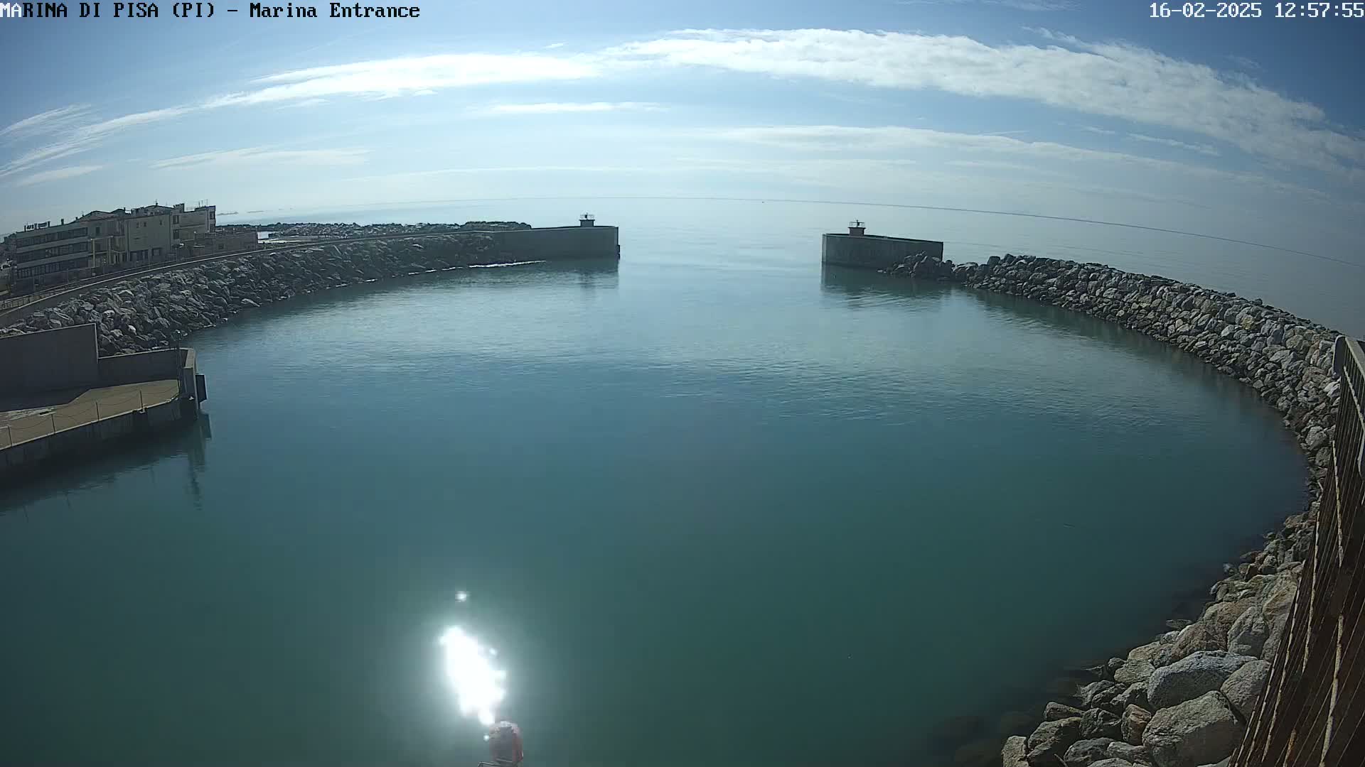 A harbor entrance with calm, teal water under a mostly sunny sky is bordered by rock walls and a building.