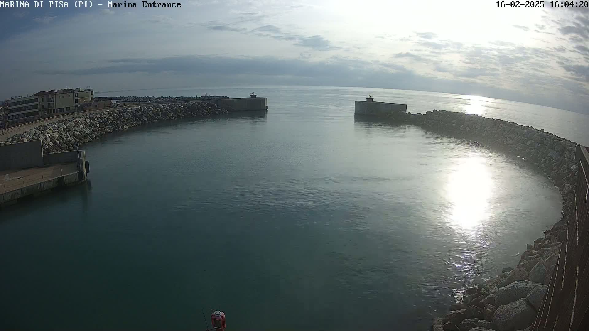 A marina entrance with calm, somewhat overcast waters is enclosed by rock walls and buildings are visible in the background.