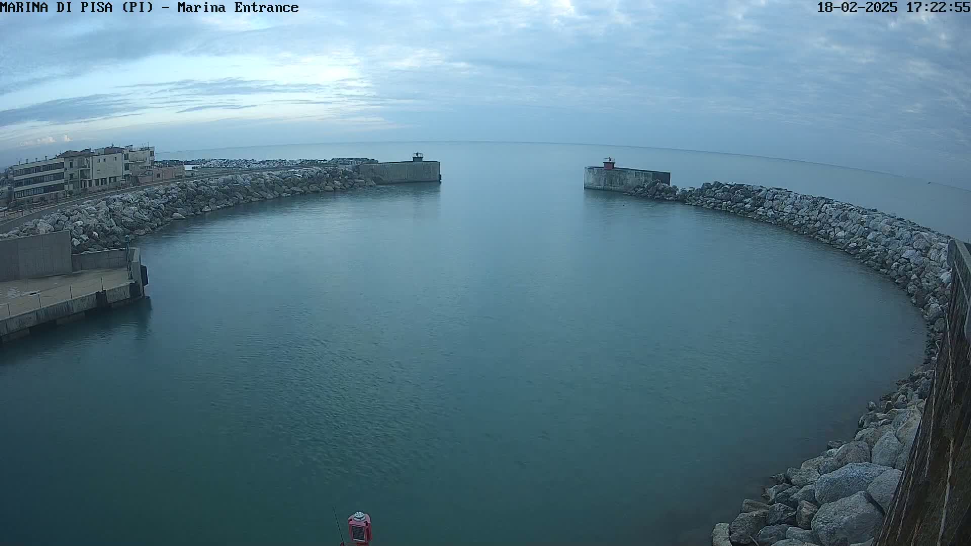 A calm, overcast day reveals a harbor entrance framed by stone walls and a building in the distance.
