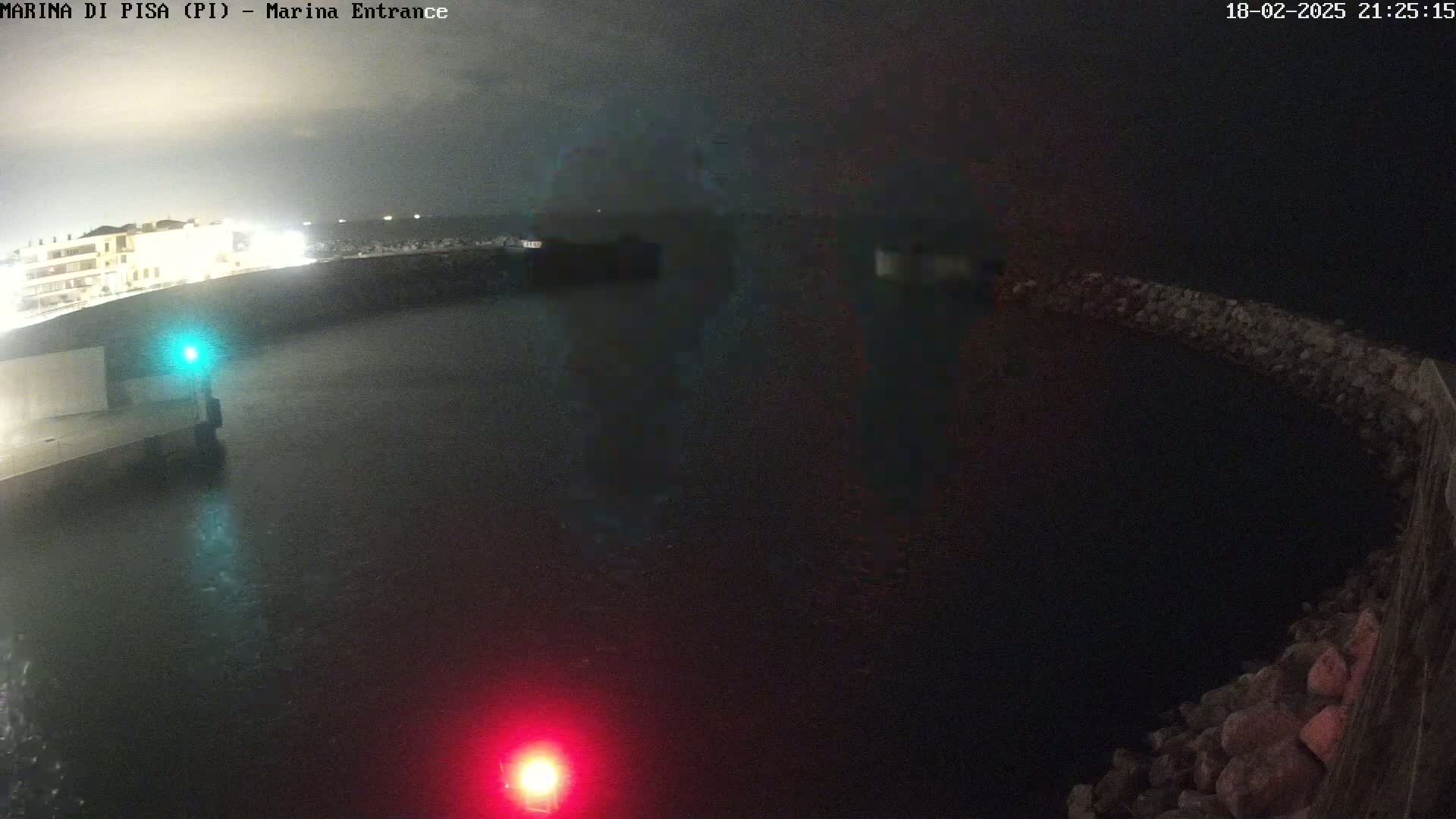 A dark, nighttime view of a harbor entrance shows calm water, a building in the distance, and stone walls on either side of the harbor.