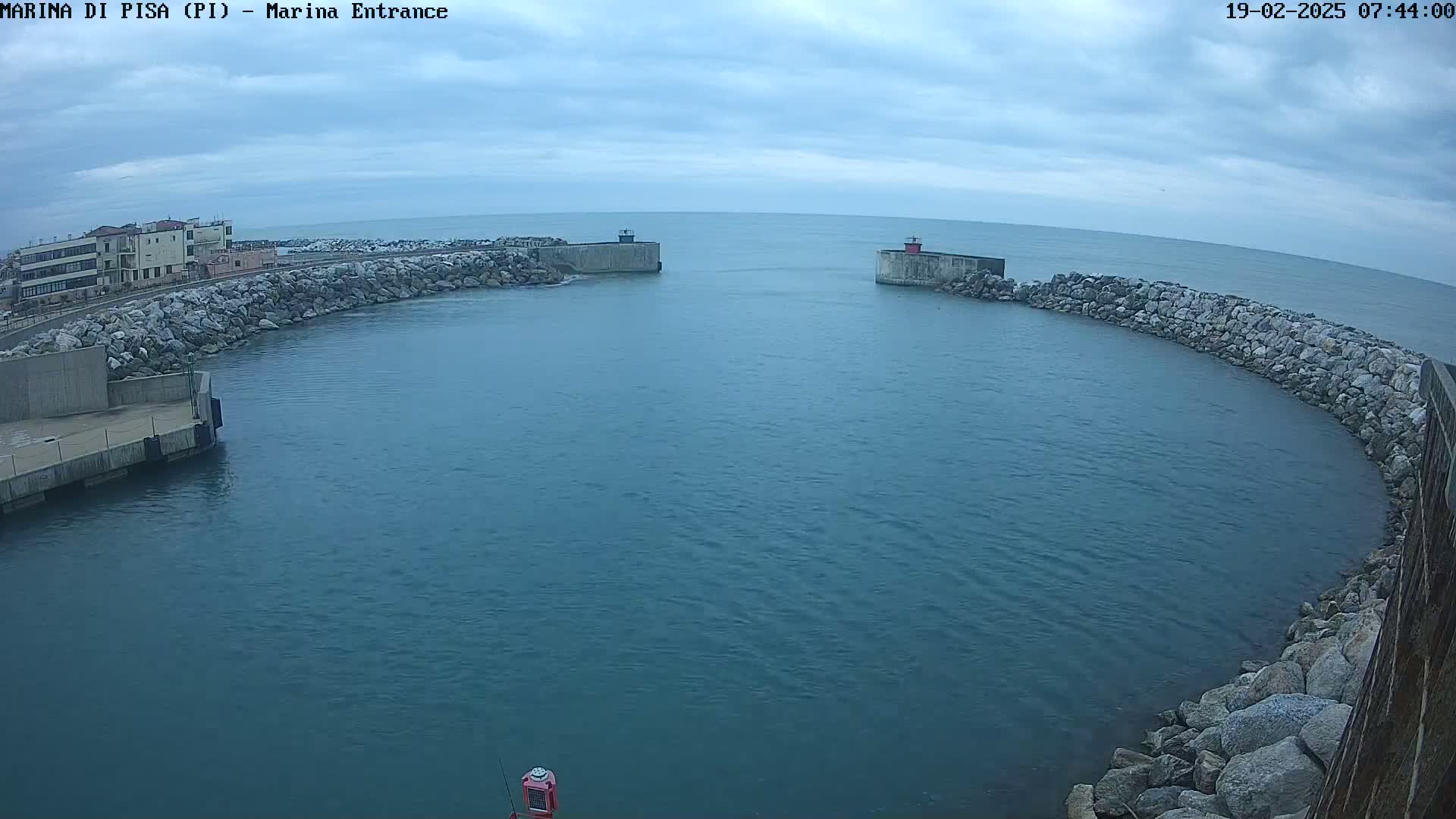 A calm, overcast day reveals a harbor entrance with two stone breakwaters curving around a tranquil body of water, buildings visible in the distance.