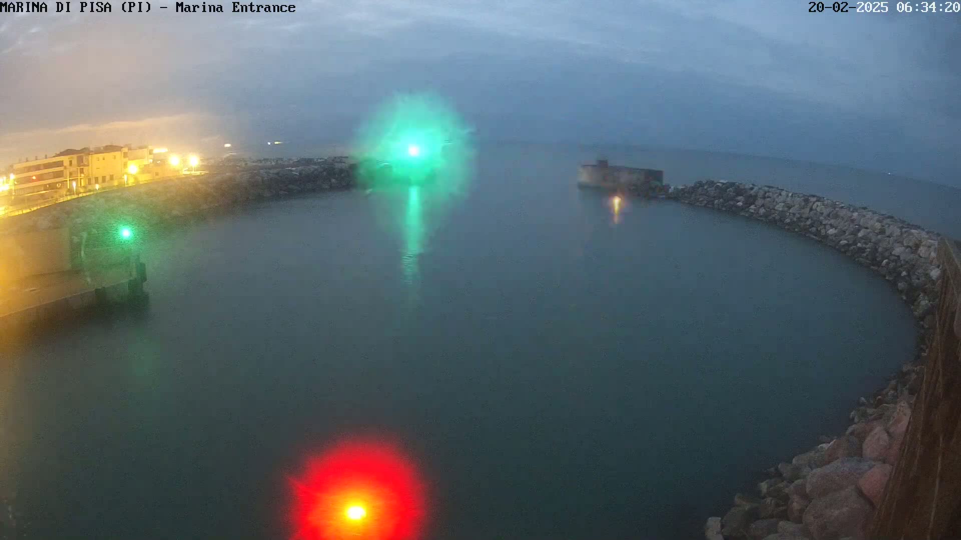 A nighttime view of a calm harbor, partially enclosed by a rock wall, with a green and a red light visible in the water.