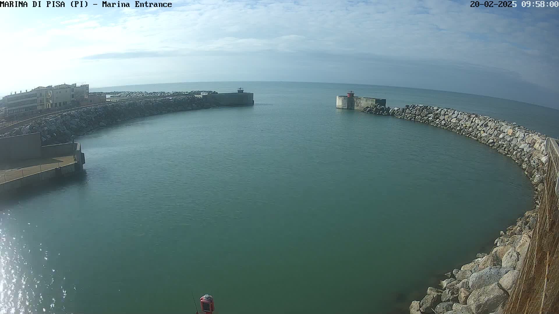 A mostly sunny day shows a calm body of green water enclosed by a rocky, curved seawall, with a small building visible in the distance.