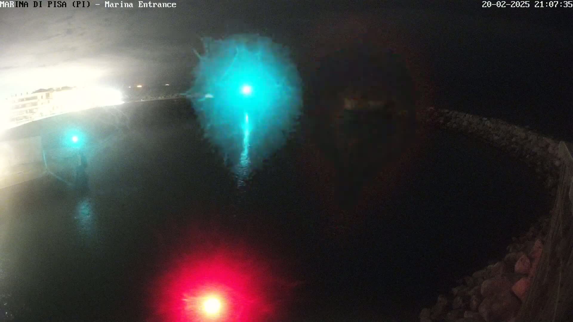 A nighttime aerial view shows a dark body of water with a bright teal light in the center and a red light at the bottom, with a rocky structure visible on the right.
