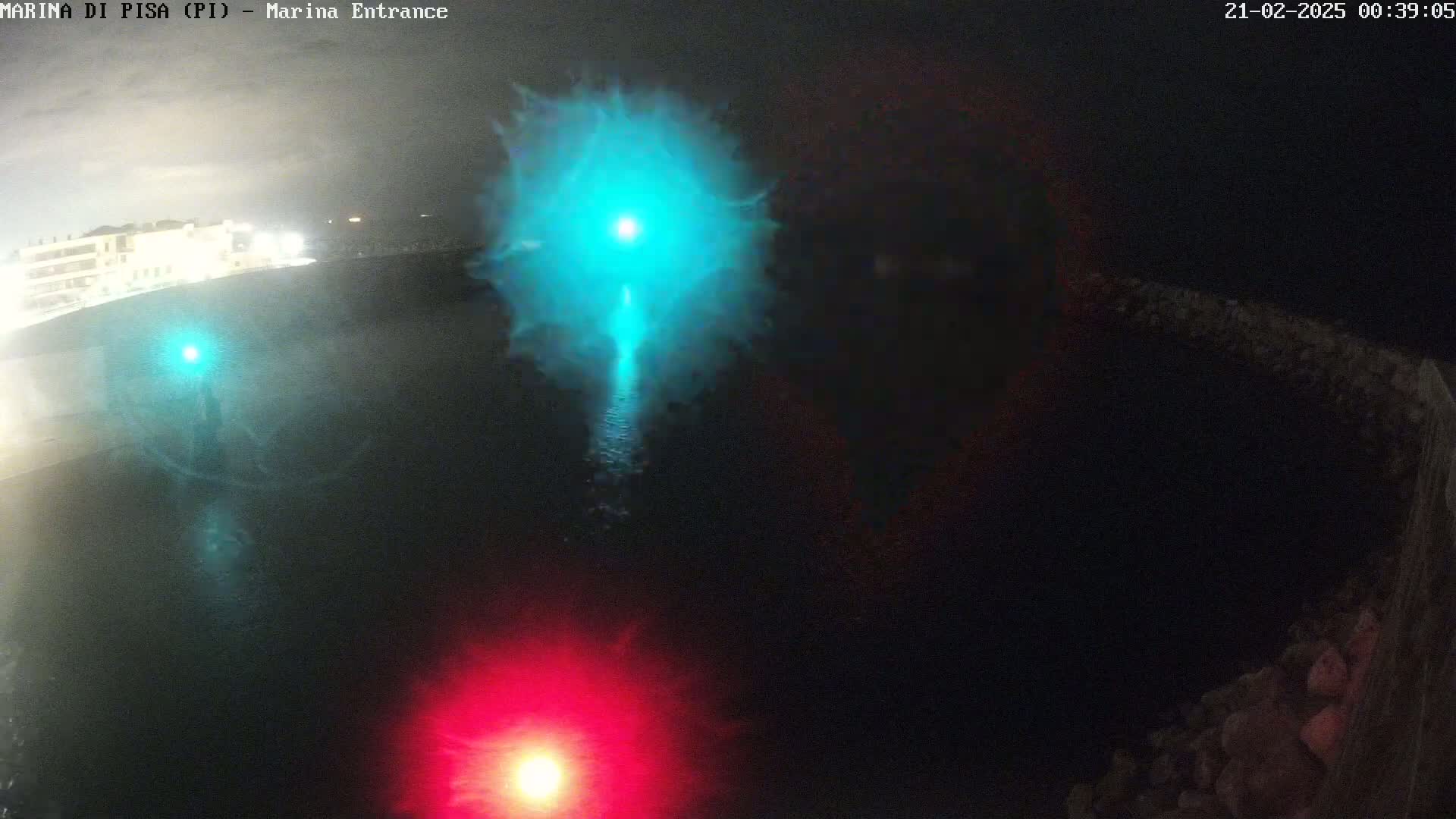 A nighttime aerial view shows a dark body of water with a bright teal light source in the center and red and green lights on either side.