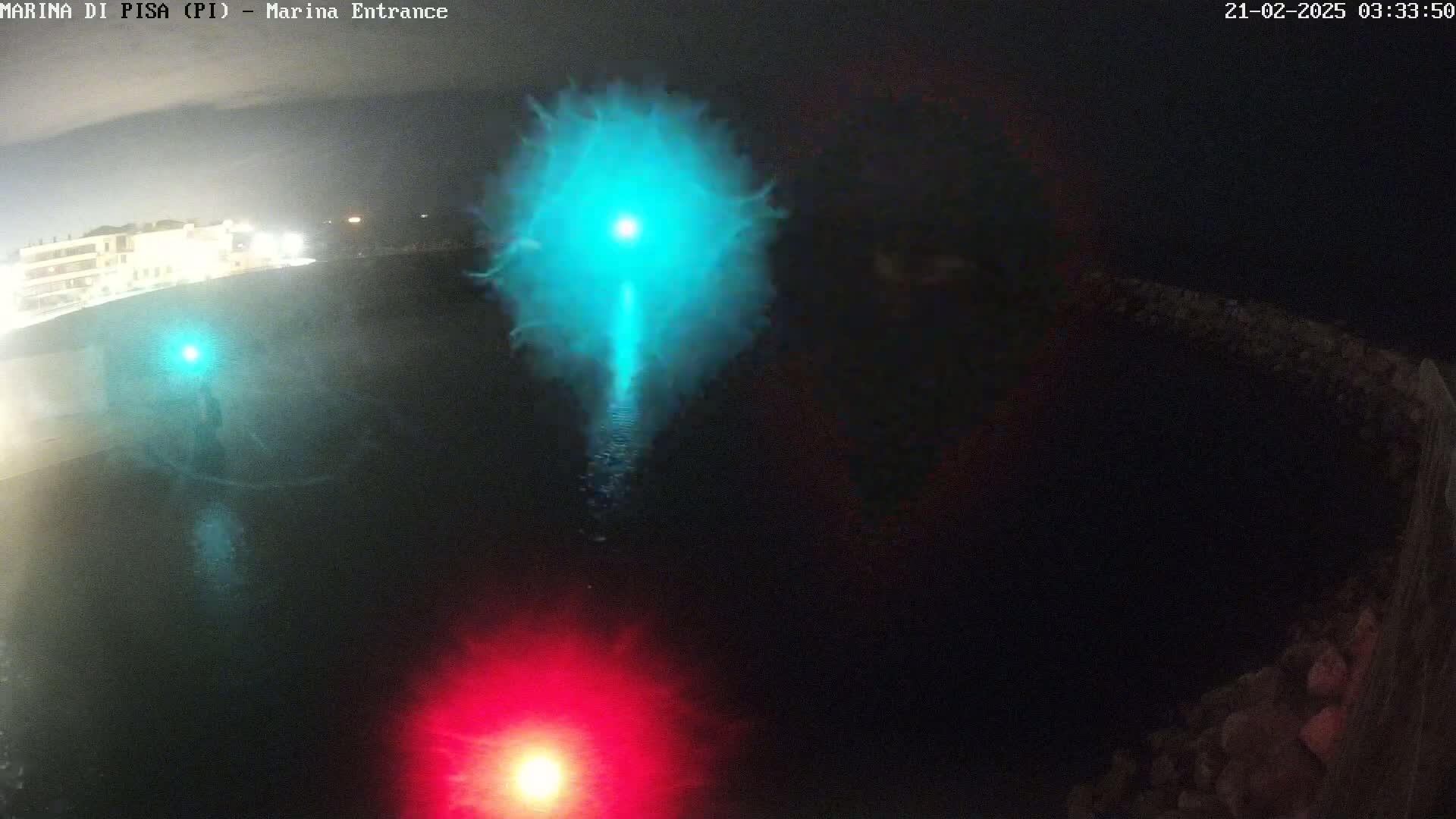 A nighttime aerial view shows a dark body of water with a bright teal light source in the center and a red light source near the bottom, illuminated against a dark shoreline.