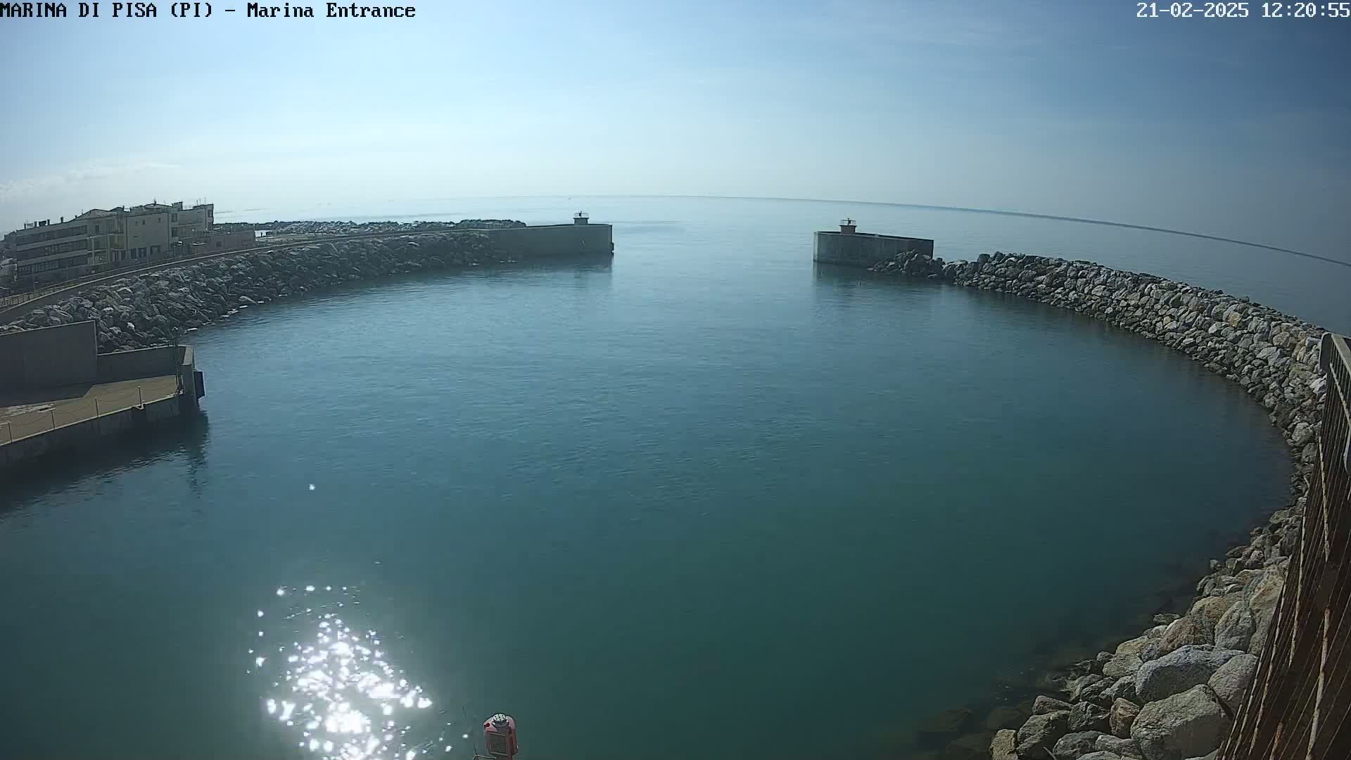 A calm, clear day reveals a harbor entrance framed by rock walls, with buildings visible in the distance.
