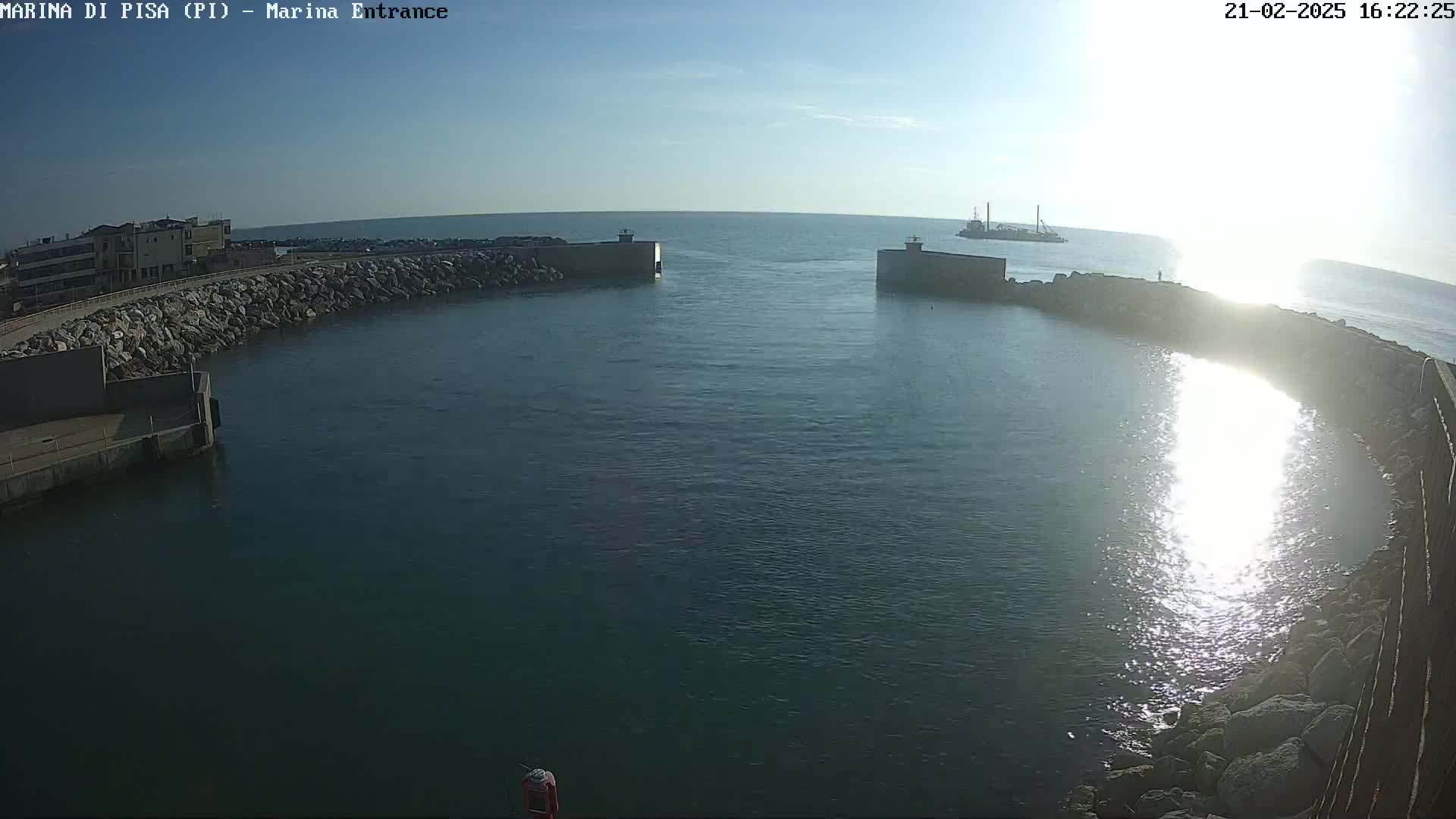 A harbor entrance with calm water reflecting sunlight, showing a distant vessel and buildings on the shore under a clear sky.