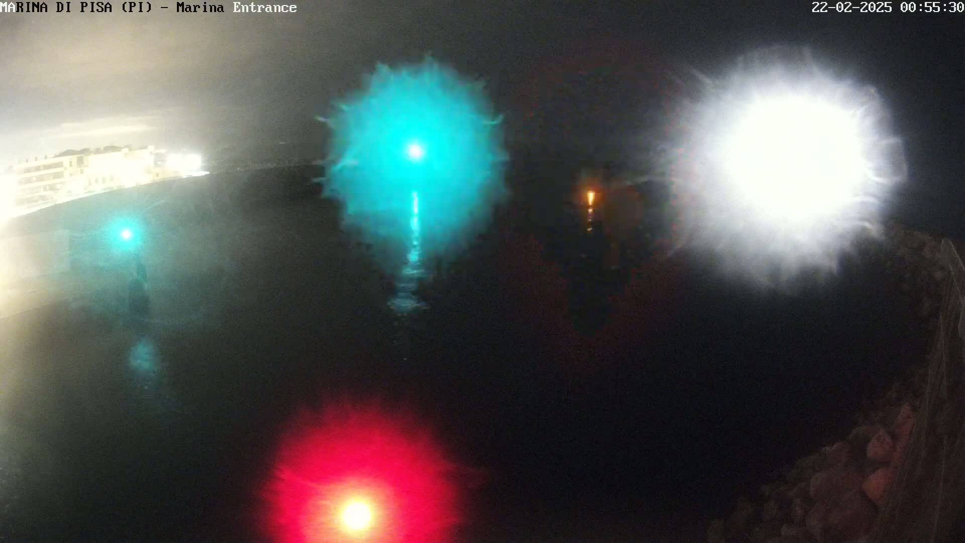 At night, bright lights of teal, white, and red illuminate a dark body of water near a shoreline.