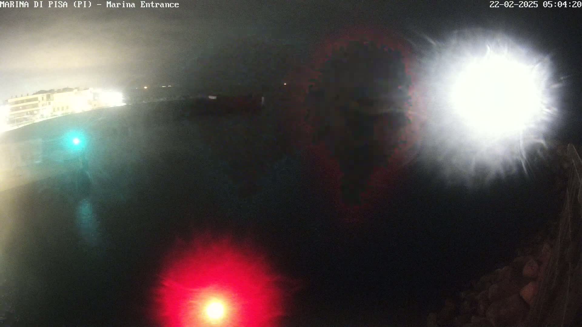 A nighttime view of a dark body of water with a bright light source in the upper right and red and teal lights in the lower portion, along with a faintly visible shoreline in the background.