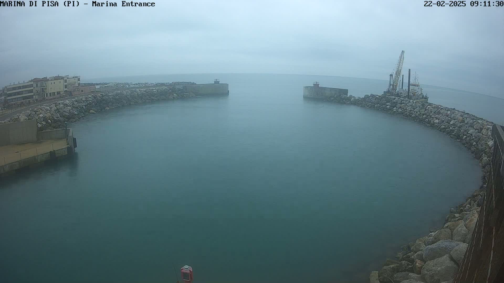 A hazy, overcast day reveals a harbor entrance with a vessel and crane at work near a rocky, curved seawall.