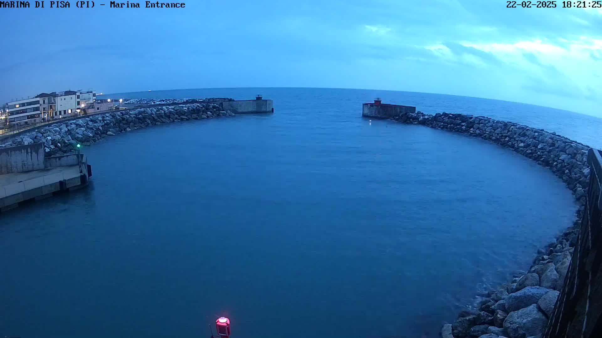 A calm, dark-blue sea is partially enclosed by a curved rock wall, with a building visible in the distance under a mostly clear, dusky sky.