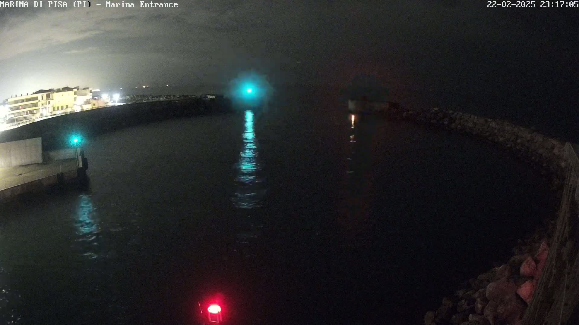 A nighttime view of a dark harbor with lights reflecting on the water, and a building visible in the background.