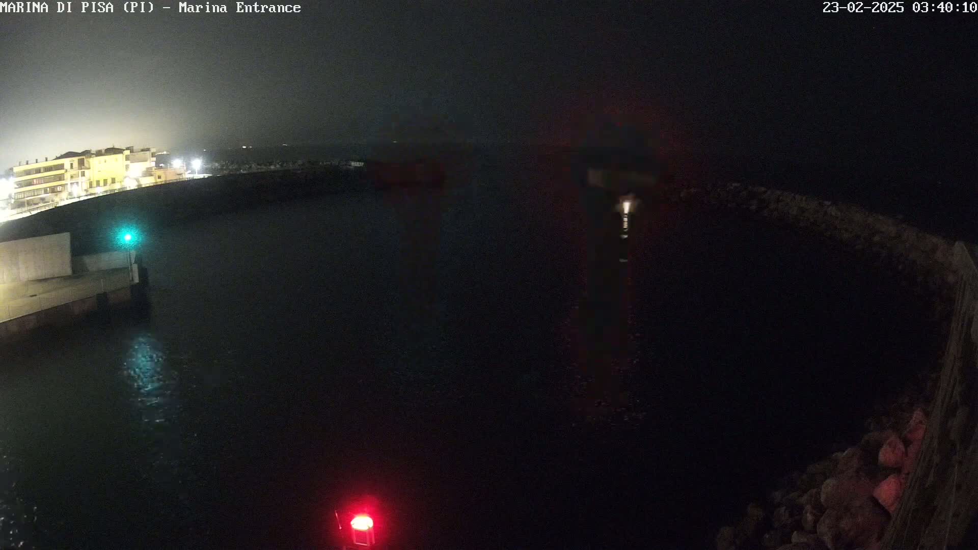 A nighttime view of a dark harbor with a building and lights faintly visible in the background, under seemingly calm water conditions.
