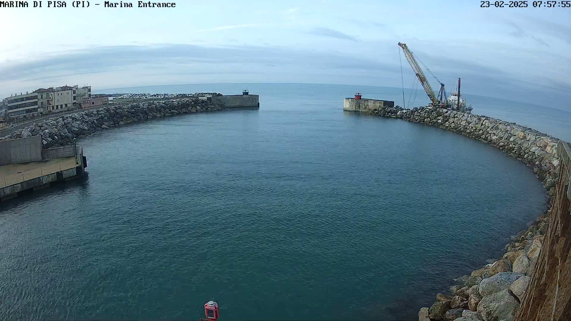 A harbor entrance with calm water, stone walls, a building in the background, and a crane on a boat.