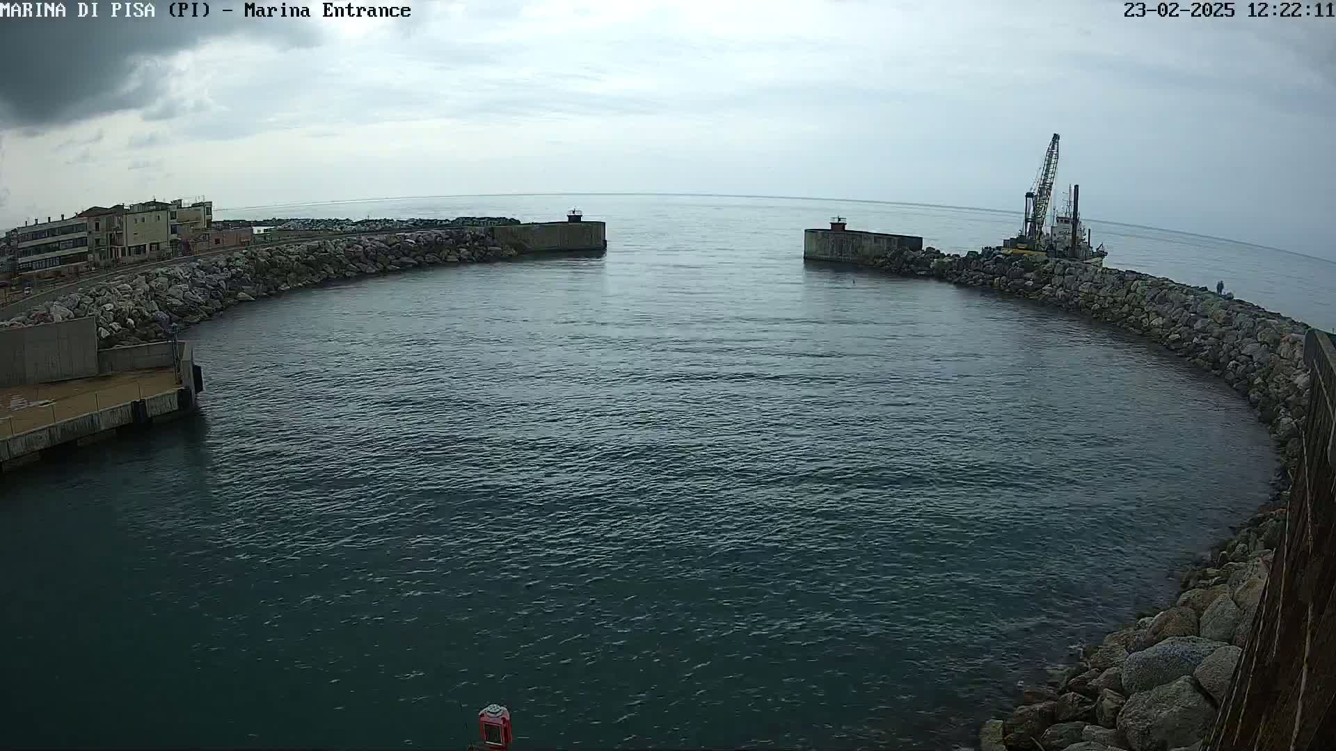 A harbor entrance with calm, dark water is partially enclosed by rocky walls, with a crane-equipped vessel visible near one of the walls under an overcast sky.