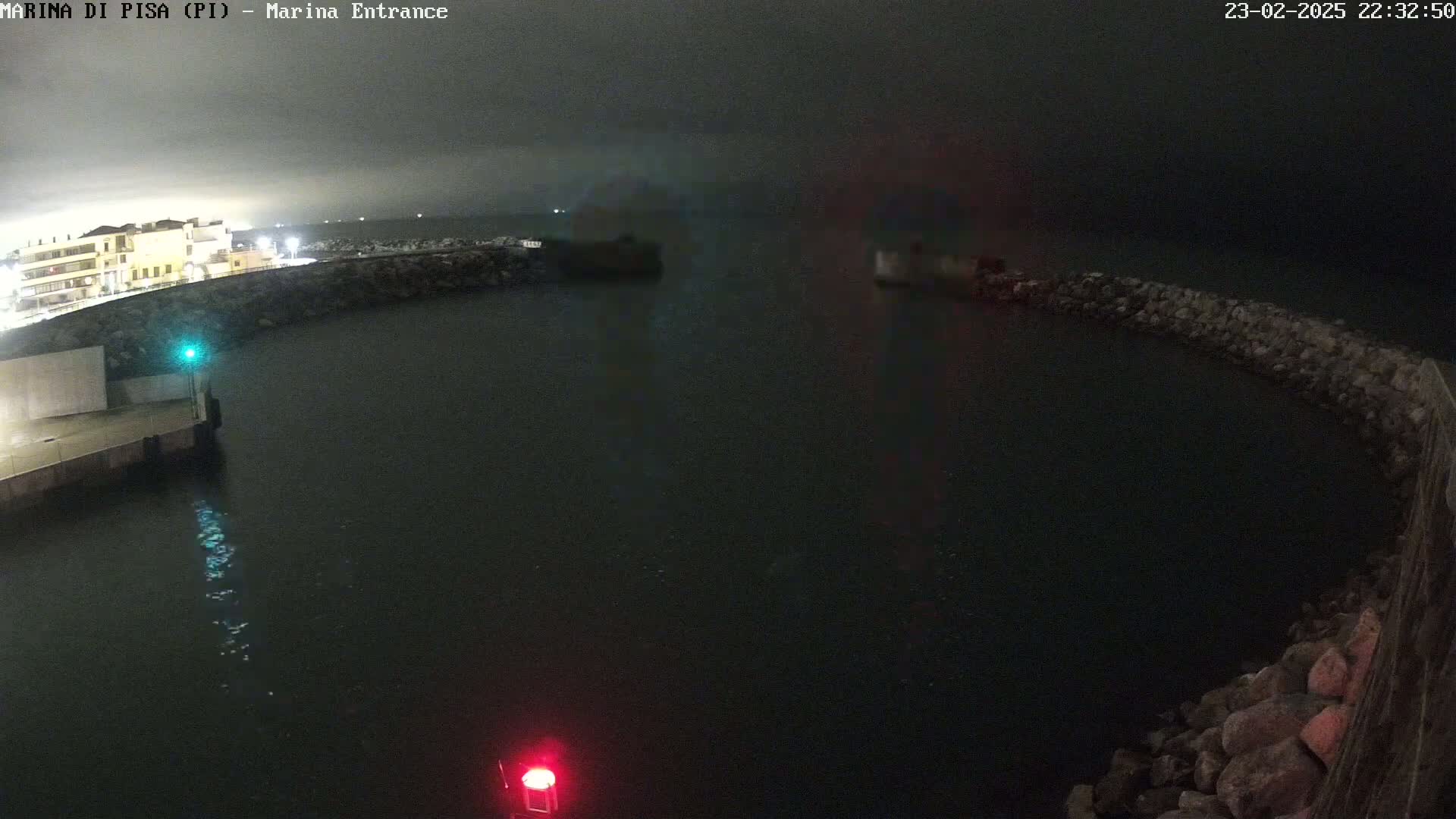 A dark nighttime view of a harbor entrance, partially enclosed by a stone wall, with calm water.