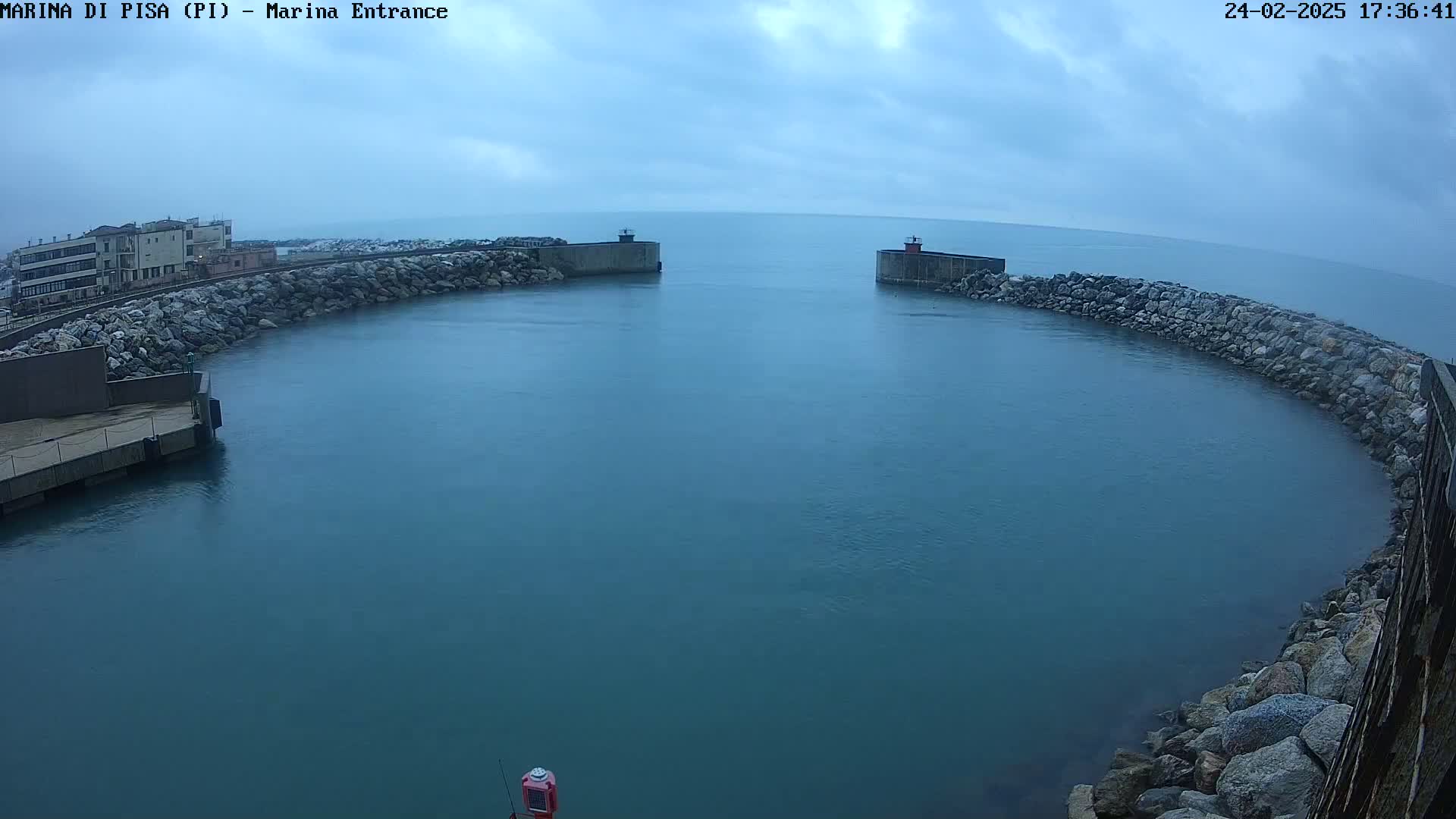 A harbor entrance with calm, dark-blue water is partially enclosed by rock walls under an overcast sky.