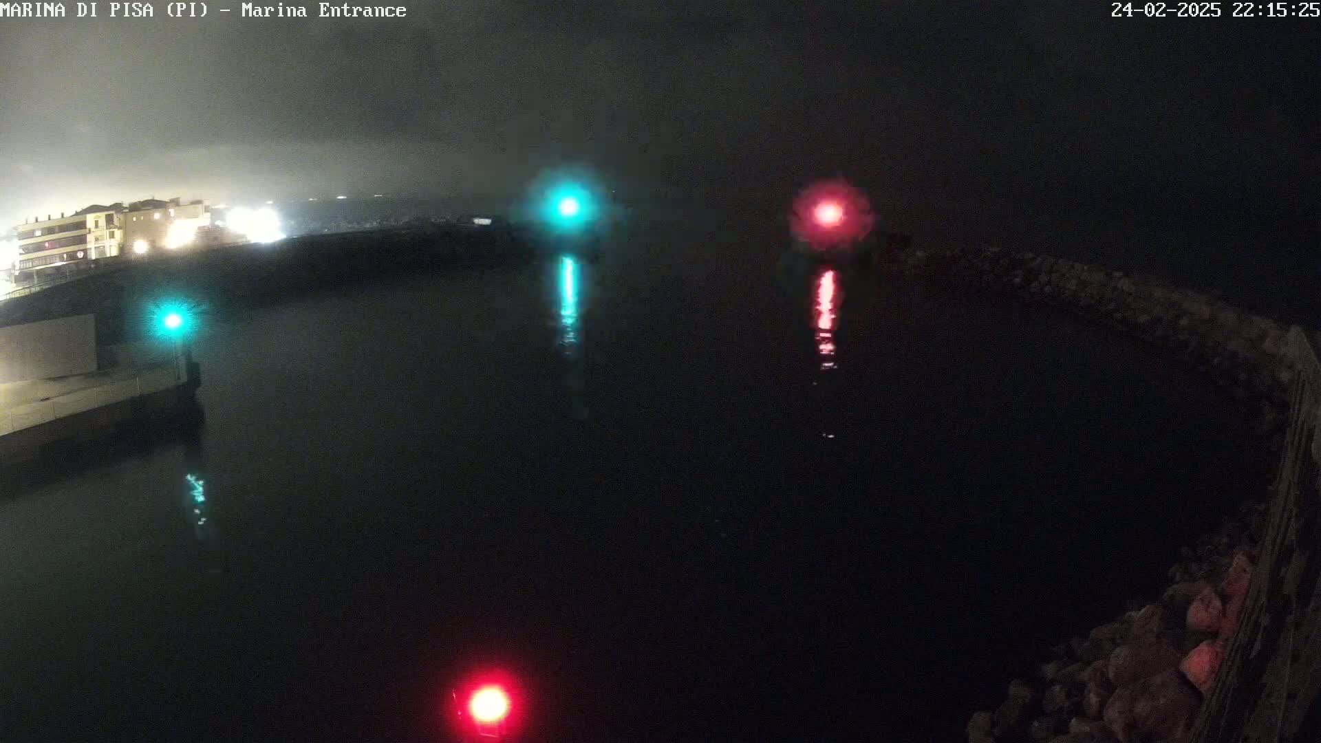 A nighttime view of a dark body of water with a red and green light reflecting on the surface, near a stone wall and a building with lights.