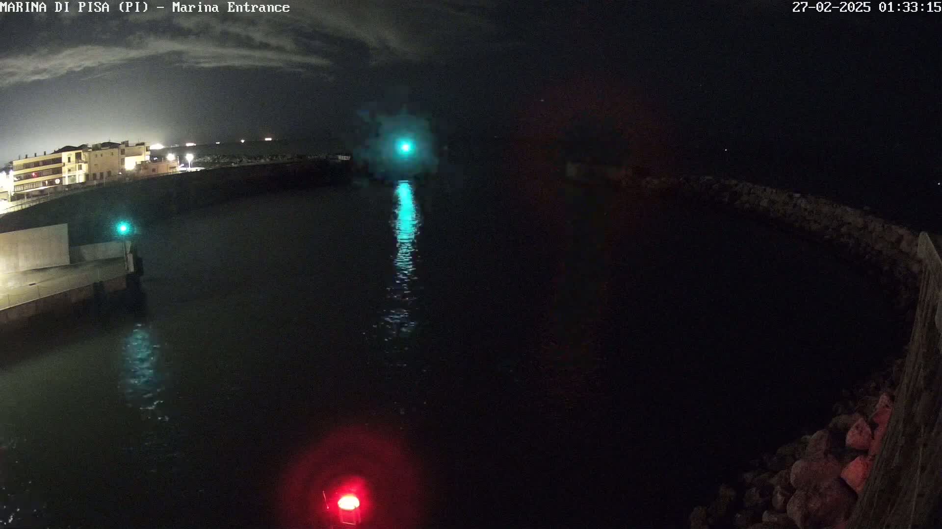 A nighttime view of a harbor entrance with a teal light reflecting on the dark water, under a mostly clear night sky.