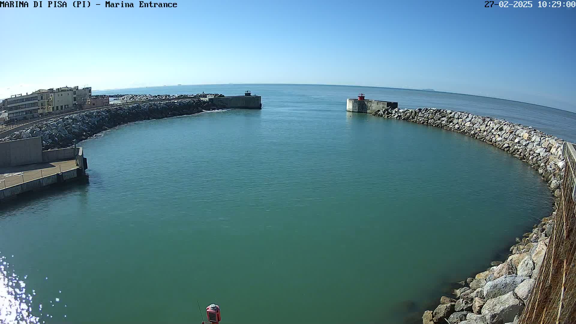 A calm, teal-colored harbor is nestled between two stone breakwaters on a sunny day.