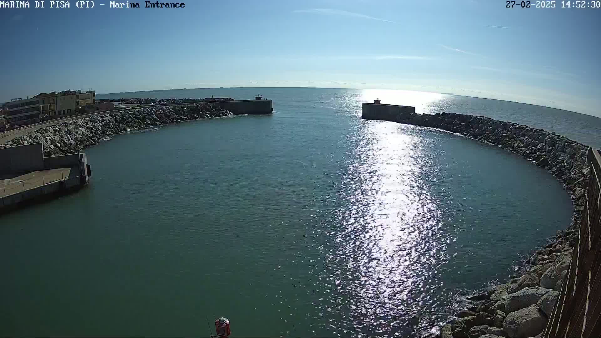A sunny day reveals a harbor entrance with calm, sparkling water partially enclosed by rocky walls and a building visible in the distance.