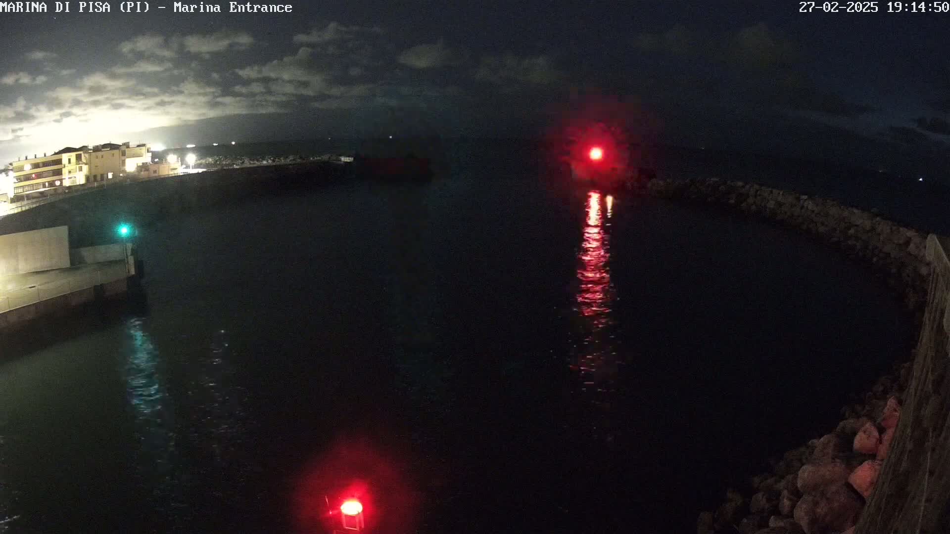 A nighttime view of a harbor entrance with calm water, illuminated by red and green navigation lights, under a partly cloudy sky.