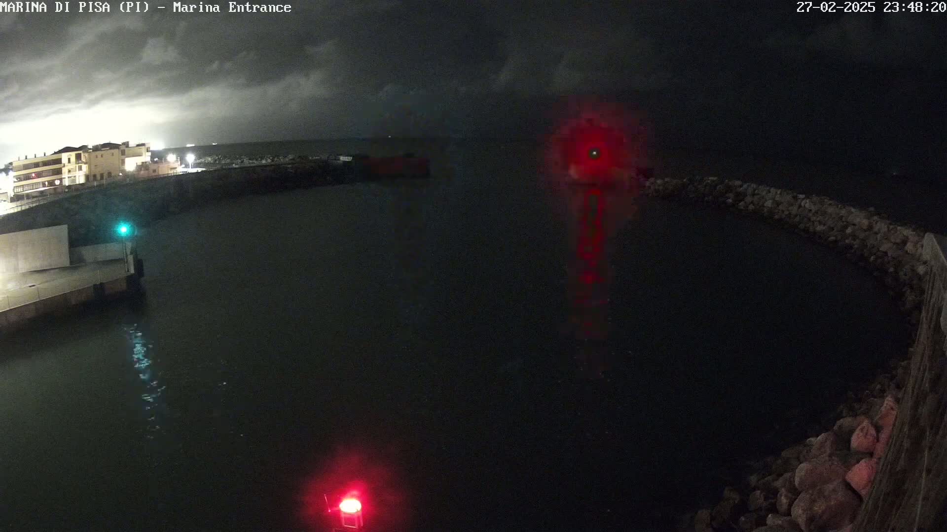A nighttime view of a dark harbor with a building in the background under a cloudy sky, illuminated by lights and harbor markers.