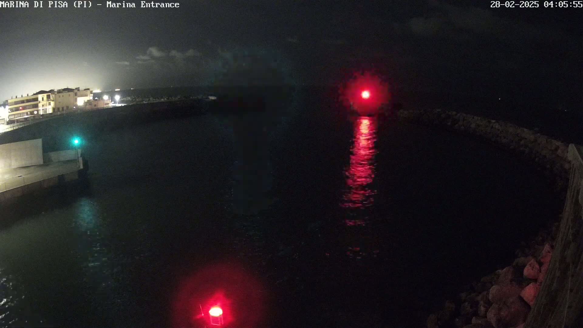 At night, red navigation lights illuminate a dark body of water next to a shoreline with a building in the background under a mostly clear sky.