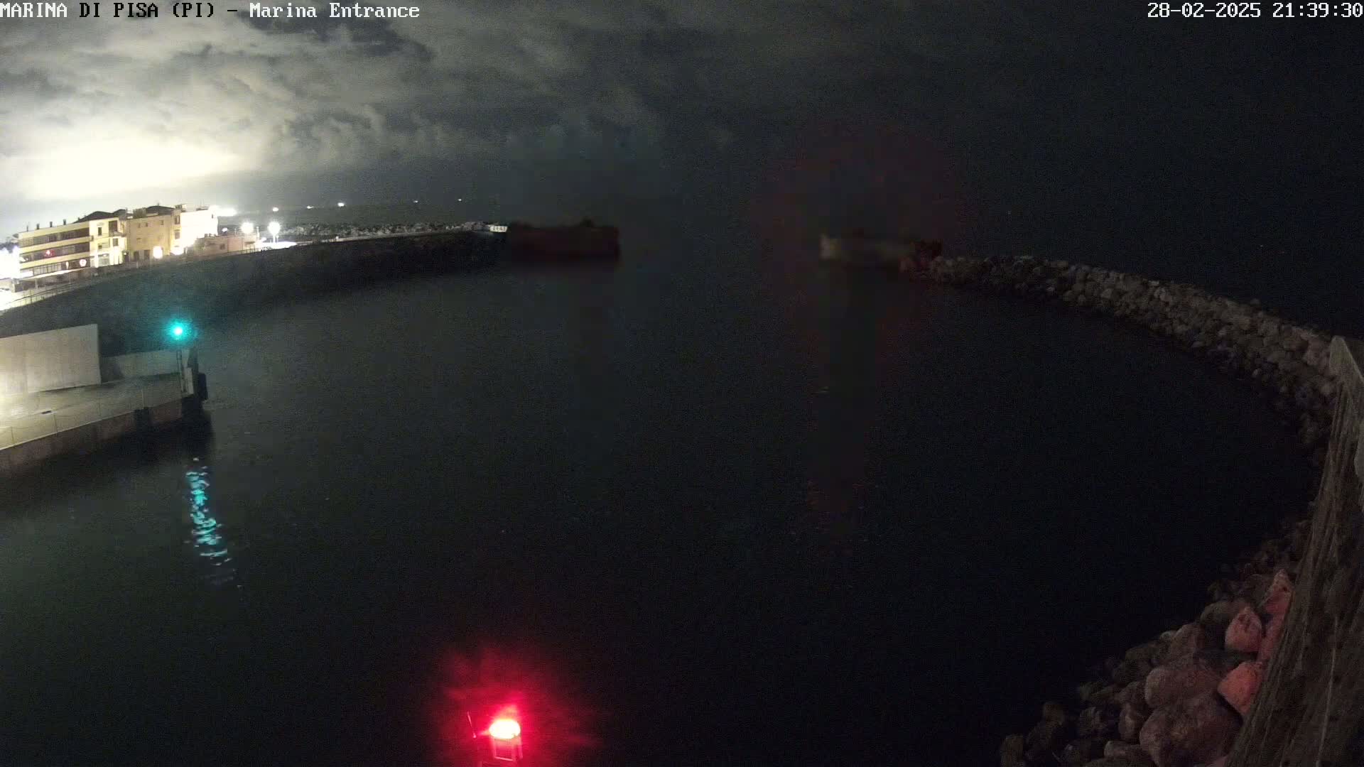 A dark nighttime scene shows a calm body of water with a stone jetty, buildings faintly visible in the background under a cloudy sky.