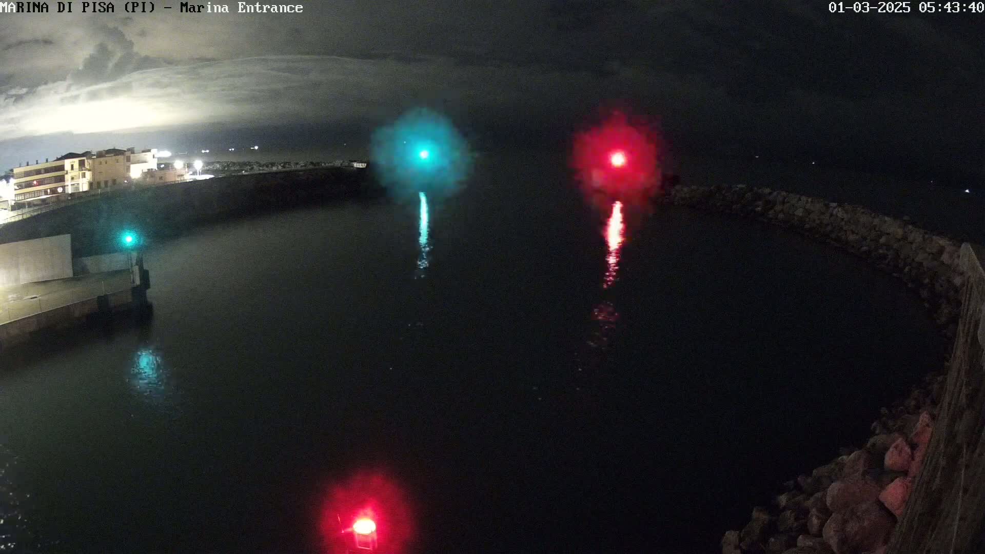 A nighttime harbor scene shows a teal and a red light emanating from the water, with a building visible in the background under a dark, cloudy sky.